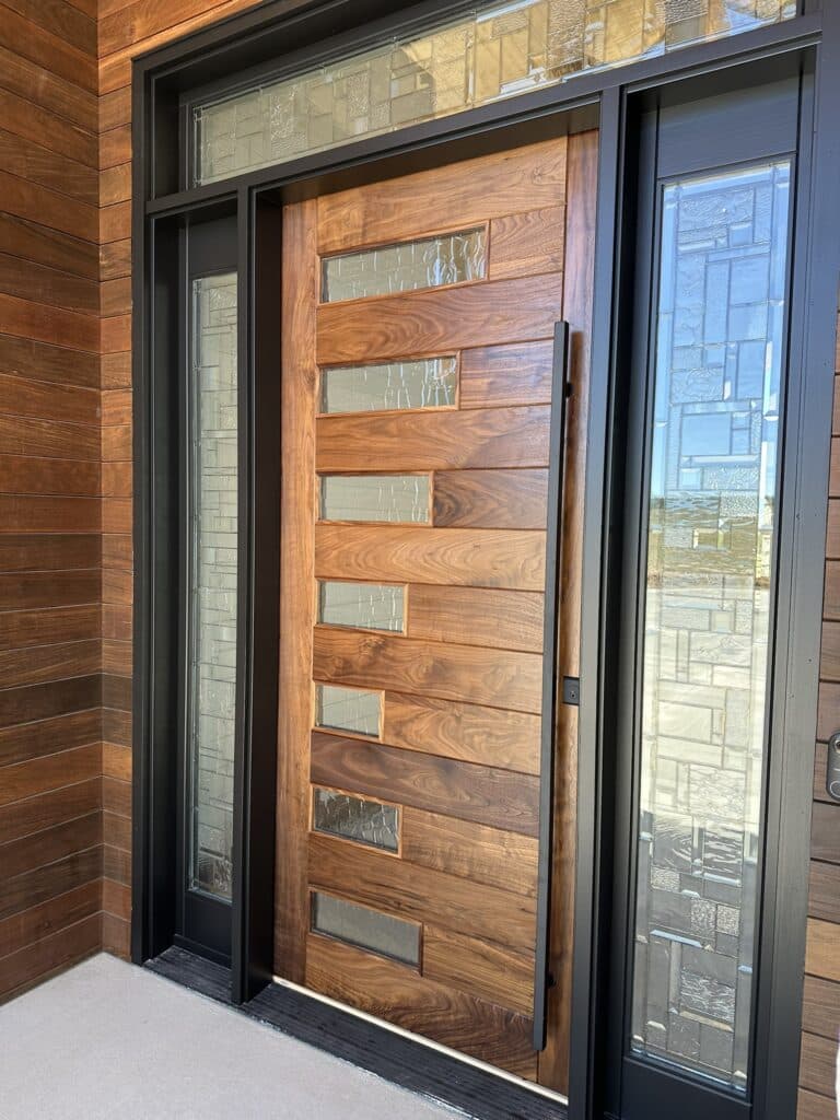 A contemporary solid wood front door with horizontal grain and rectangular clear textured glass inserts, part of an entryway system featuring black-framed sidelights and a transom window, all fitted with clear geometric leaded glass panels. The surrounding exterior wall is clad in horizontal wood planks.