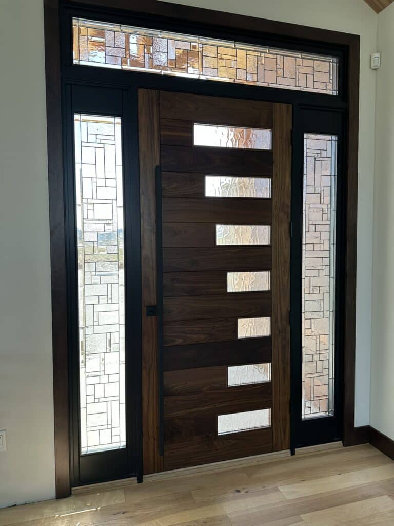 A dark wood modern door with rectangular privacy glass inserts, flanked by stained glass sidelights and topped with a stained glass transom, all featuring clear, textured linear geometric patterns.