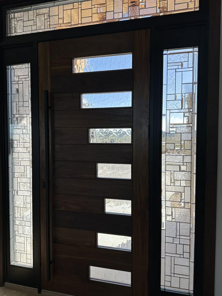 A contemporary entryway featuring a dark wood door with multiple horizontal clear textured glass inserts, flanked by matching leaded glass sidelights and a transom above, all displaying an abstract geometric pattern with clear textured and beveled glass pieces.