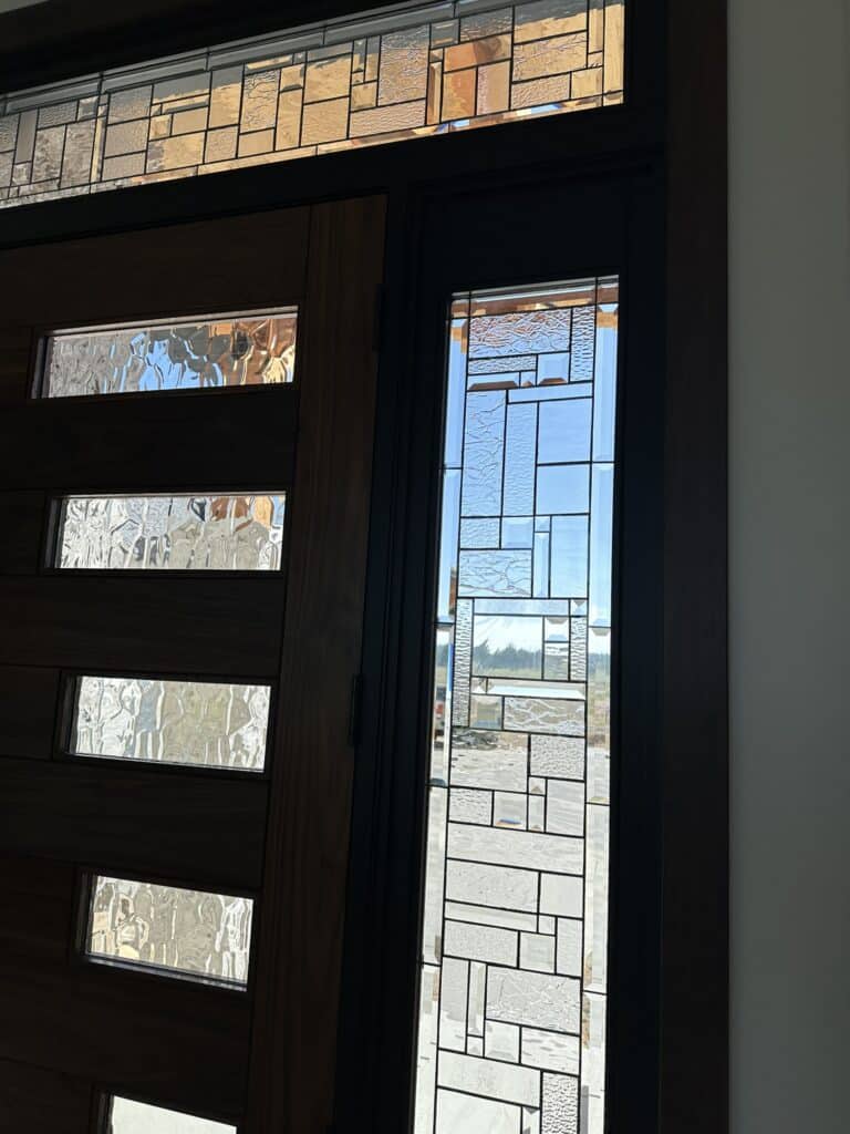 A contemporary entry door system featuring a dark wood door with horizontal clear textured glass inserts, alongside a clear leaded glass sidelight and transom, both exhibiting an abstract geometric pattern with various textured and bevelled glass pieces.