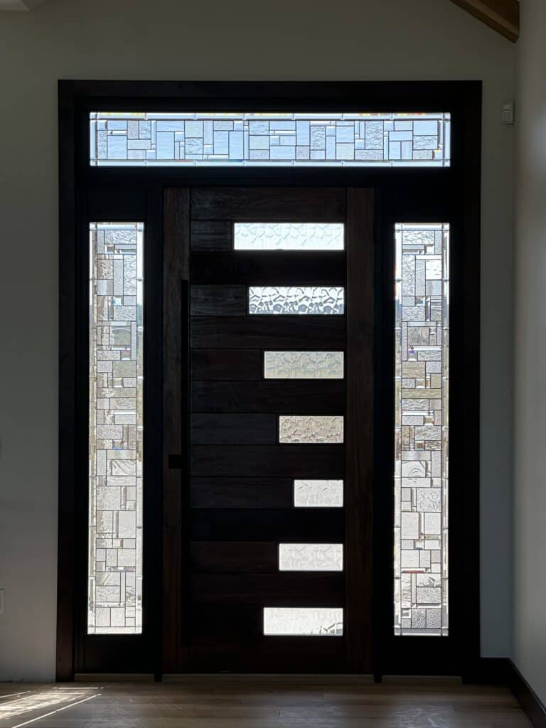 A modern dark wood entry door with multiple rectangular clear glass inserts, flanked by matching abstract geometric clear stained glass sidelights and topped with a coordinating transom window.