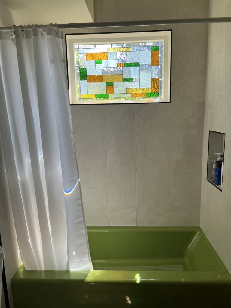 A rectangular stained glass window with an abstract geometric pattern in clear, amber, and green textured glass, installed above a green bathtub.