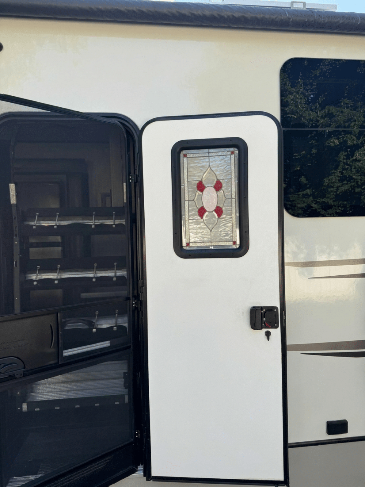 A rectangular stained glass panel with a central geometric design featuring red and clear ovals surrounded by textured clear glass, installed in a white RV door.