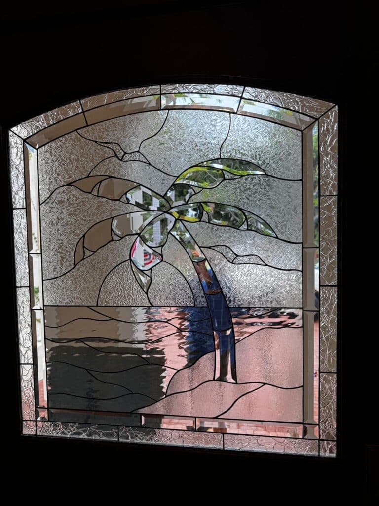 An arched, leaded stained glass panel depicting a tropical scene with a palm tree, water, and sand, made with clear textured and beveled glass.