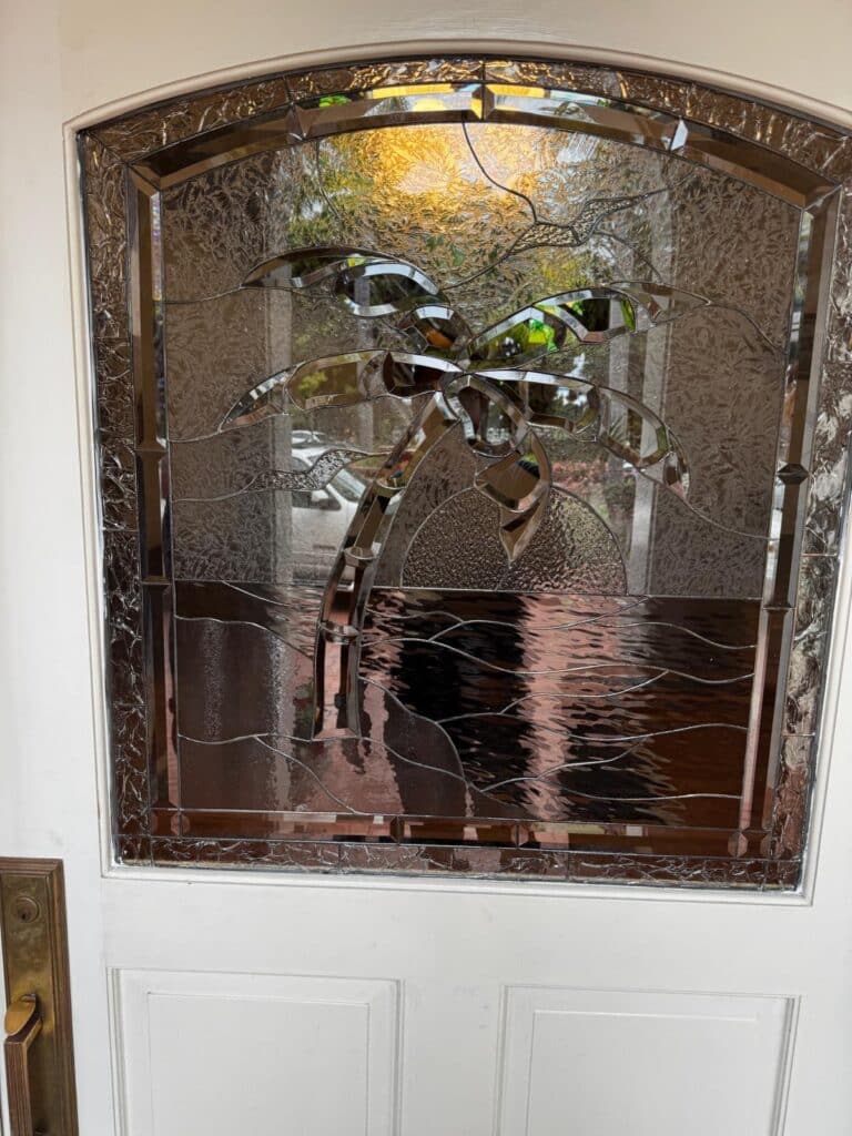 An arched, clear textured stained glass door insert depicting a tropical scene with a palm tree, setting sun, and ocean waves, all made with various types of clear and beveled glass.