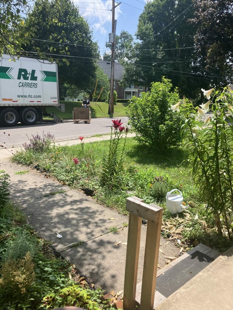 A delivery truck on a residential street, with a worker unloading a large box labeled 'STAINED GLASS' from the truck's liftgate onto a pallet jack.