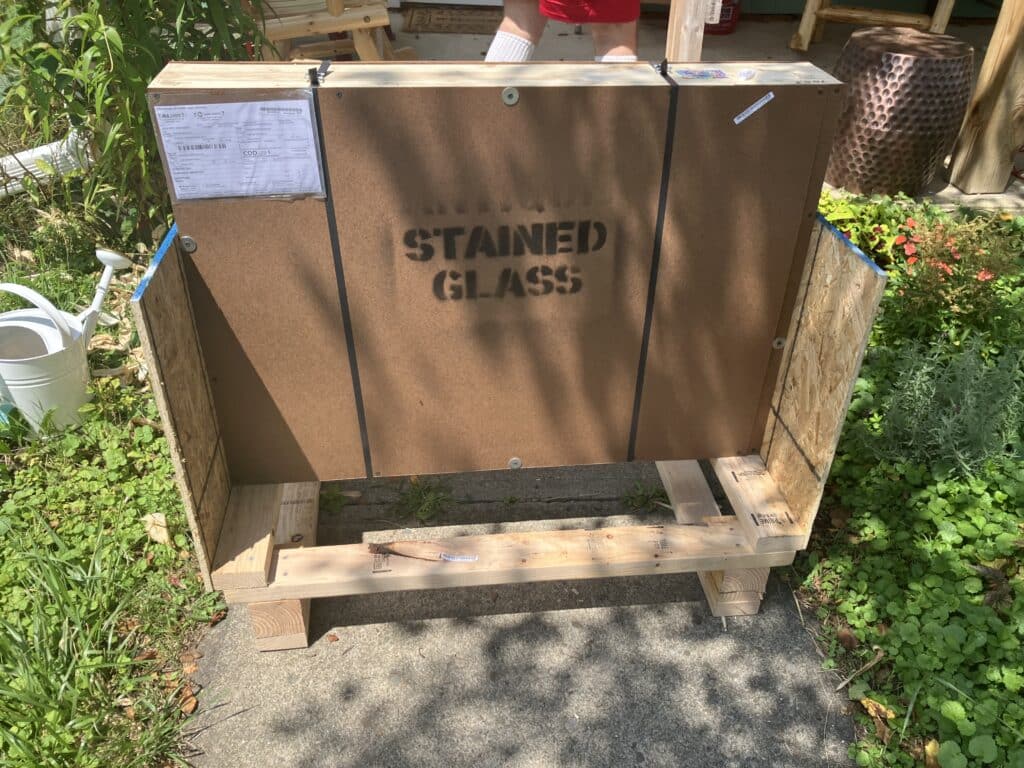 A robust wooden shipping crate, clearly labeled 'STAINED GLASS,' resting outdoors on a concrete surface, surrounded by green plants.