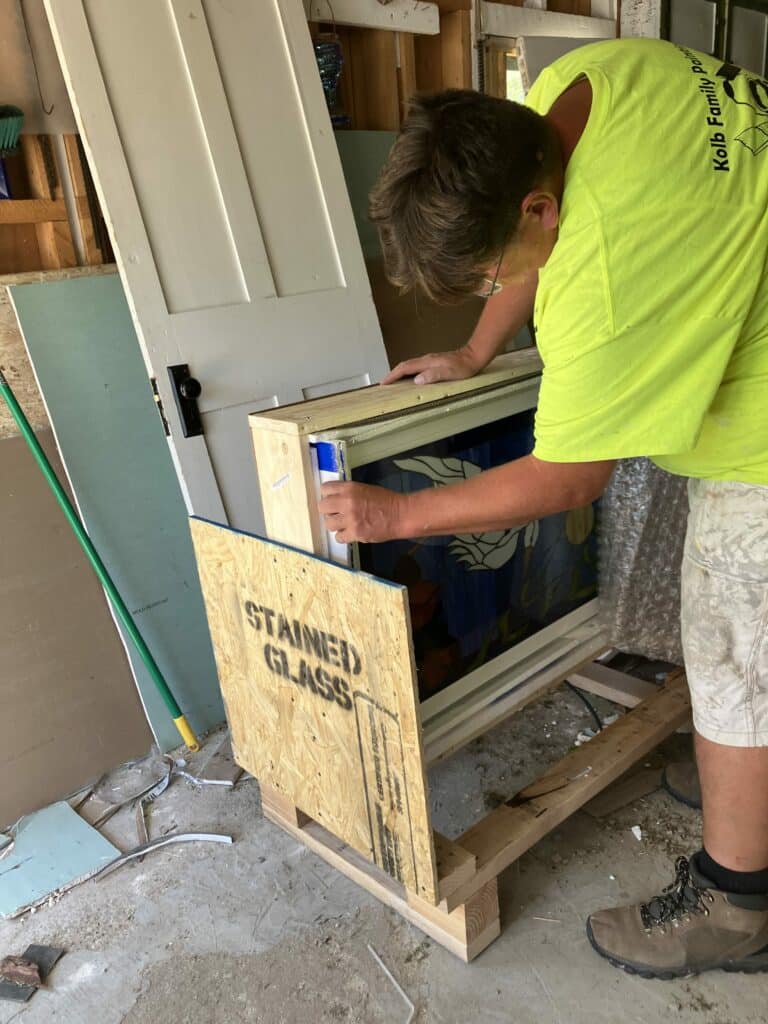 A person wearing a bright yellow t-shirt and work shorts carefully lifts a custom stained glass panel, with a white and blue abstract design, from its protective wooden shipping crate marked 'STAINED GLASS' in a workshop.