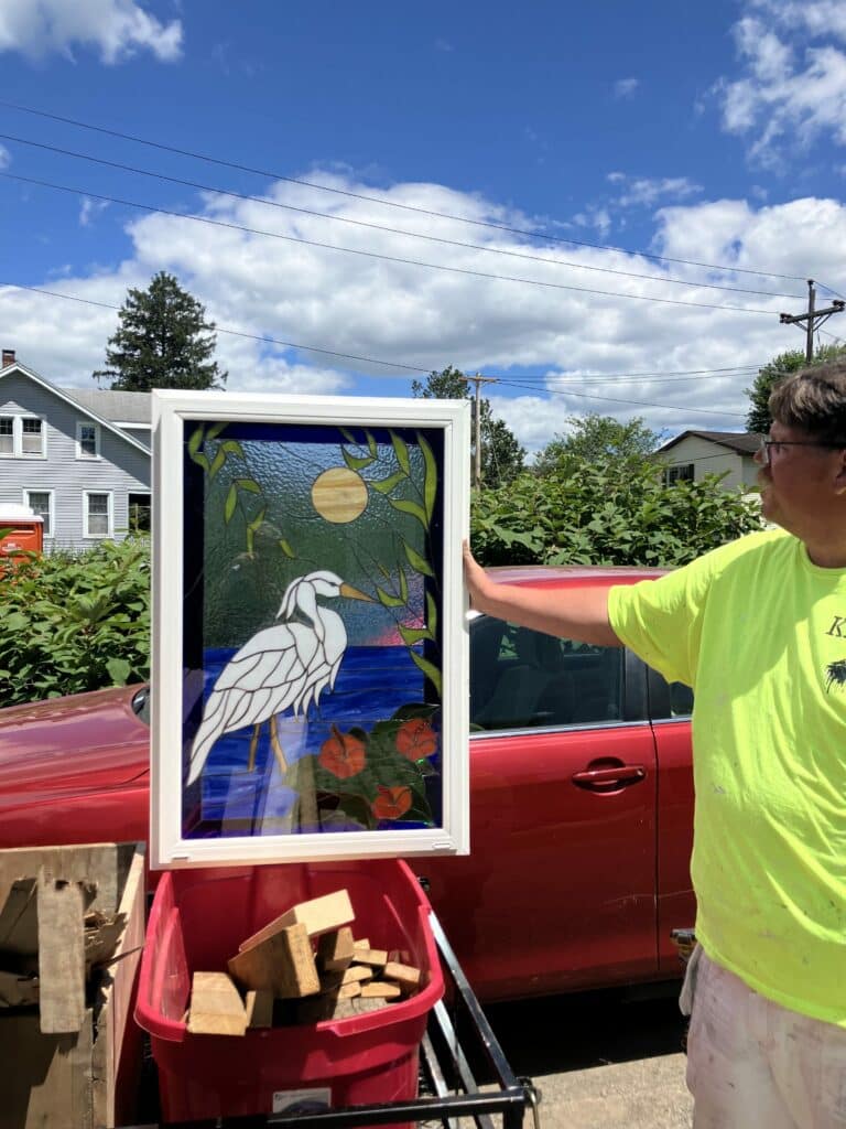 A rectangular stained glass panel in a white frame featuring a white great egret or heron standing in blue water, with a yellow full moon, green leaves, and orange hibiscus flowers.
