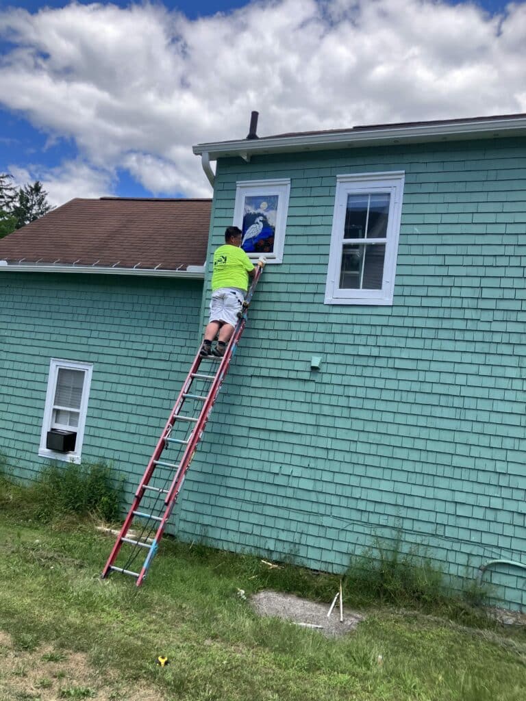 A person on a ladder installs a rectangular stained glass window panel featuring a white heron, moon, and water onto a teal-shingled house.