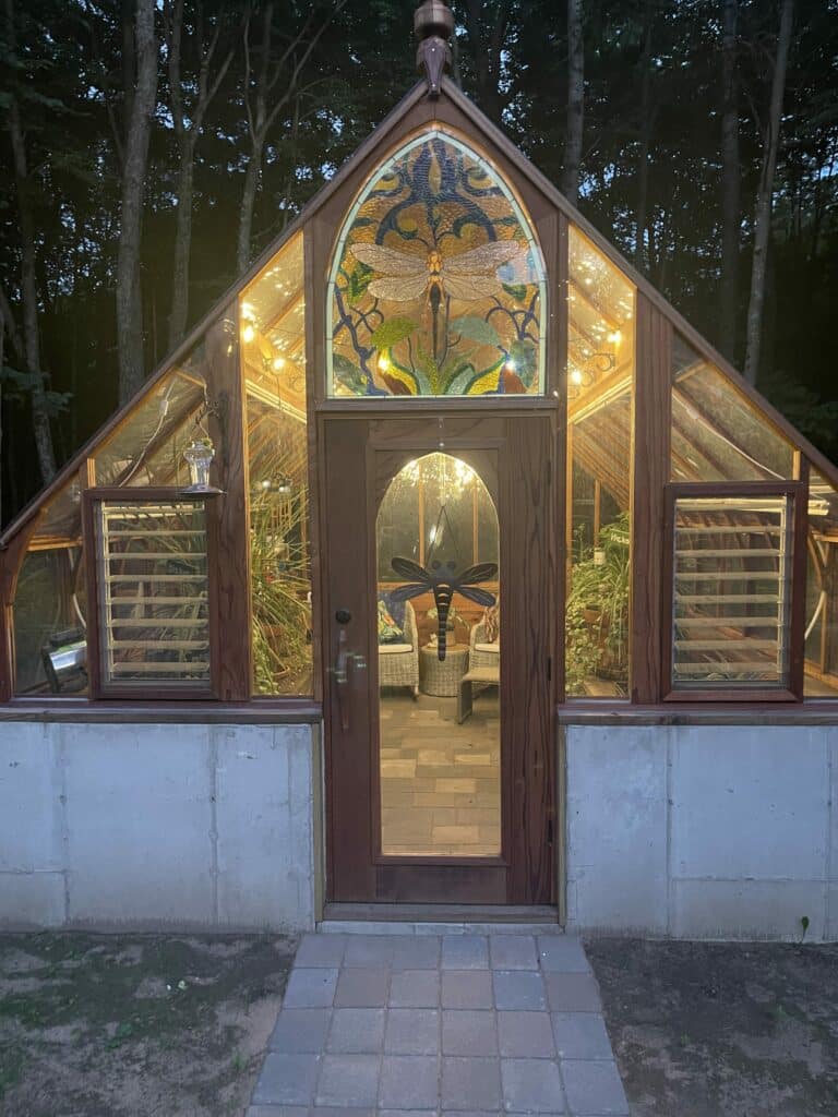 An arched stained glass panel featuring a detailed dragonfly with clear wings and colorful botanical elements, set in the gable of a wooden greenhouse, illuminated at night.