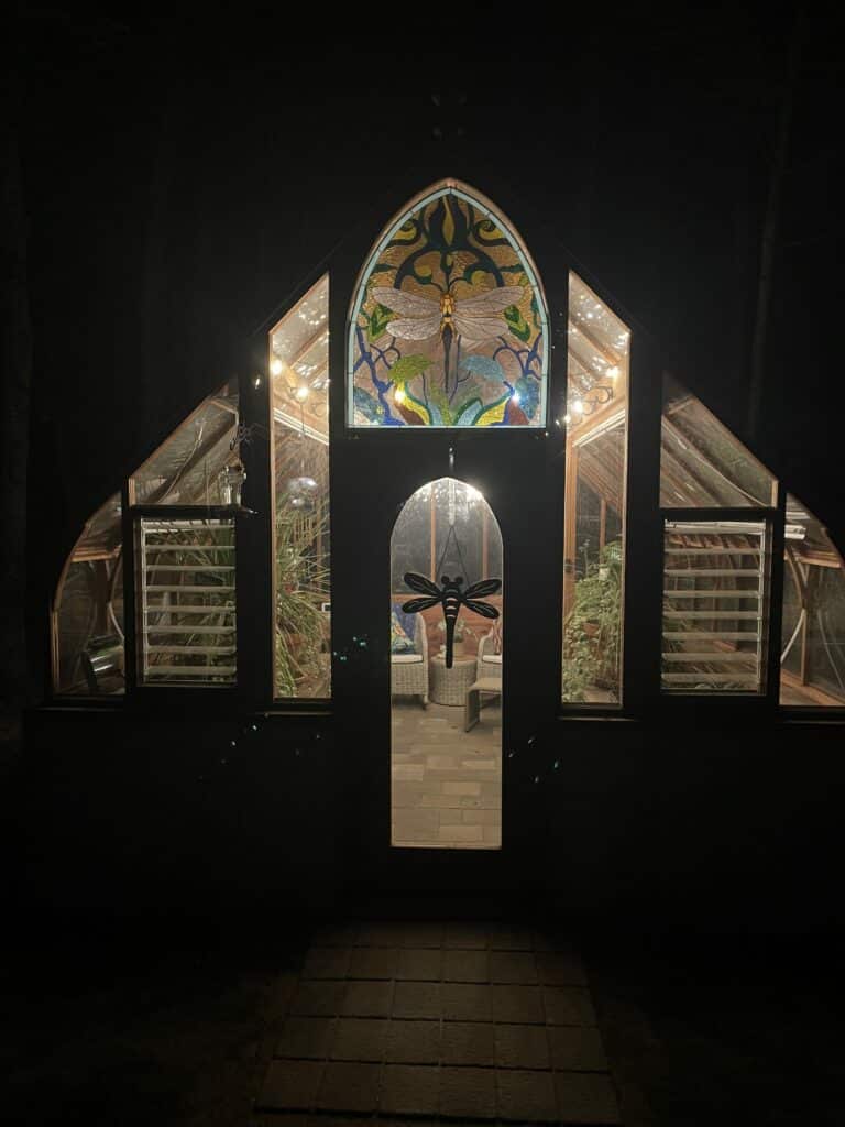 A grand arched greenhouse illuminated at night, featuring a vibrant stained glass panel at its peak. The panel depicts a large yellow and clear textured dragonfly surrounded by green and blue abstract foliage. Below, the arched glass door reveals a dark silhouette of a dragonfly hanging, leading into a brightly lit interior with wicker furniture and lush plants. The greenhouse's surrounding glass panels glow with internal string lights, set against the dark night.