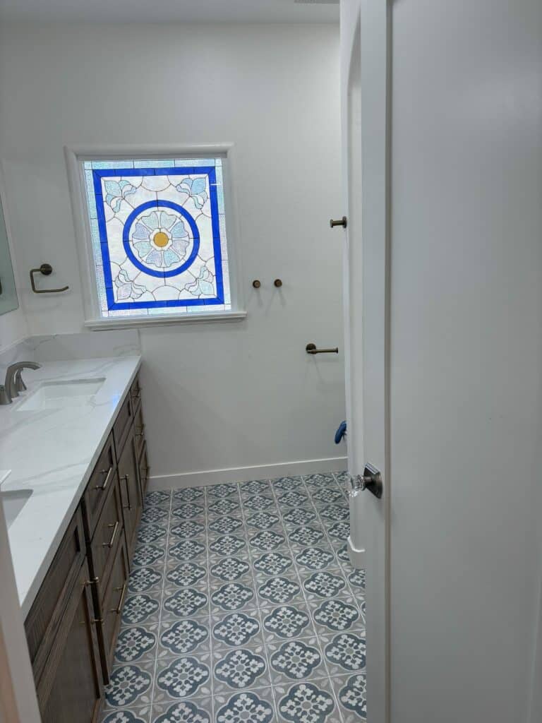 A rectangular stained glass window with a central white and blue floral medallion, bordered by a solid blue frame, set in a white bathroom.