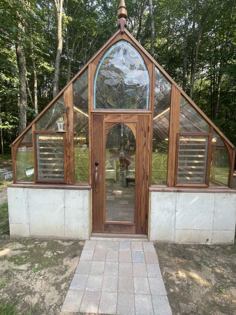 A rustic wooden greenhouse with a prominent arched stained glass panel above the door, featuring a large, clear-winged dragonfly amidst green leaves and branches. The greenhouse has a concrete foundation, louvered side windows, and a paved walkway leading to its entrance, surrounded by tall trees.
