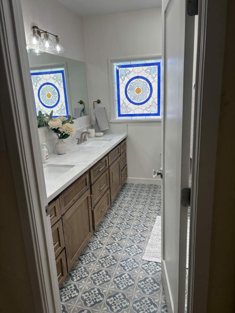 A rectangular stained glass window with a central blue and yellow floral medallion, surrounded by light blue and clear textured glass patterns, set in a bathroom.