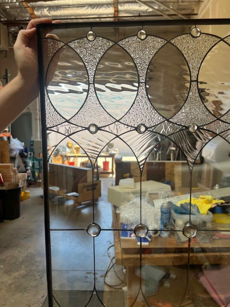A rectangular leaded stained glass panel featuring a repeating geometric pattern of clear smooth and clear textured glass circles and ovals, accented with round clear jewels. A hand holds the top left edge of the panel.