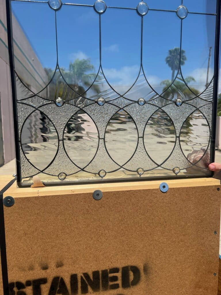 A rectangular leaded stained glass panel featuring a geometric pattern of interlocking clear ovals and textured glass sections, accented with clear round jewels. The panel is seen against a bright blue sky with a palm tree visible through the clear glass.