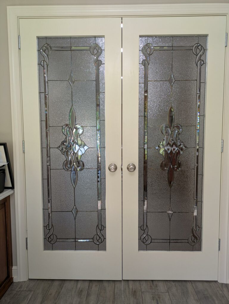A pair of white interior French doors, each featuring a rectangular leaded stained glass panel with clear textured glass and an ornate beveled fleur-de-lis motif.