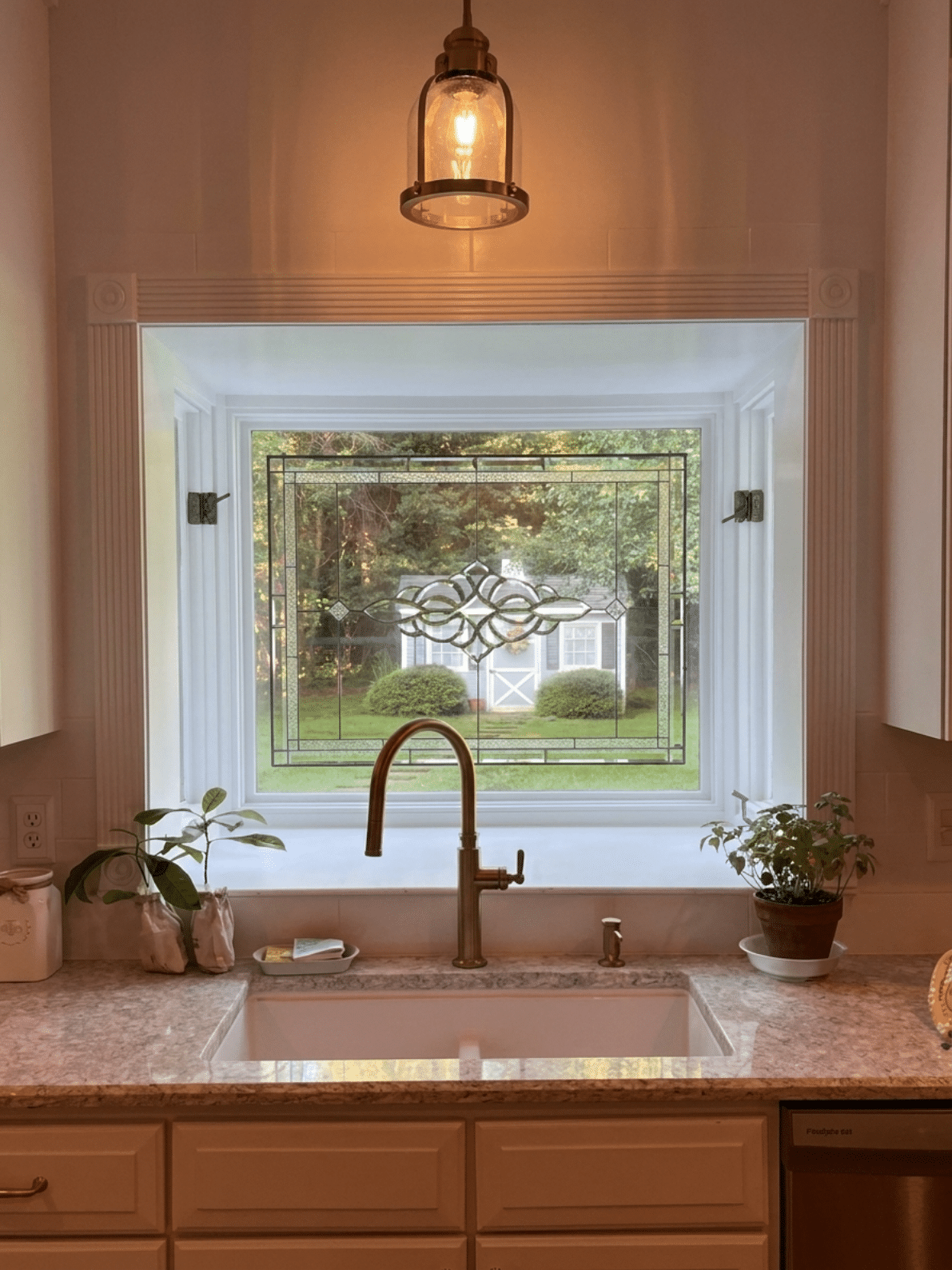A rectangular, leaded stained glass panel installed in a kitchen window above a sink. The panel features a clear textured and beveled glass border surrounding a central abstract scrollwork or organic medallion design, also in clear textured glass. The window looks out to a garden with a small shed.