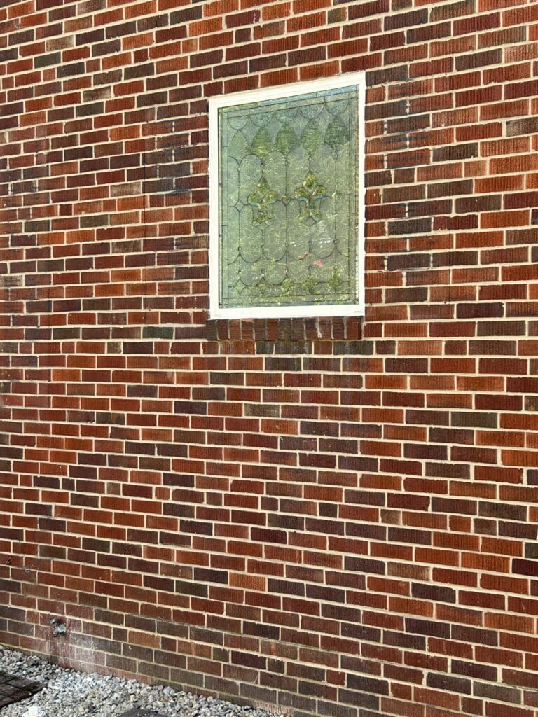 A rectangular stained glass window with a white frame, featuring two prominent green fleur-de-lis motifs, surrounded by textured clear and light green glass in geometric and scalloped patterns, set into a multi-toned red brick wall.