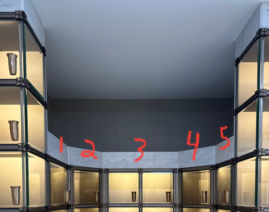 A wall of glass-fronted display niches forming a curved alcove, each illuminated from within and containing a small urn, with ornate dark metal corner details.