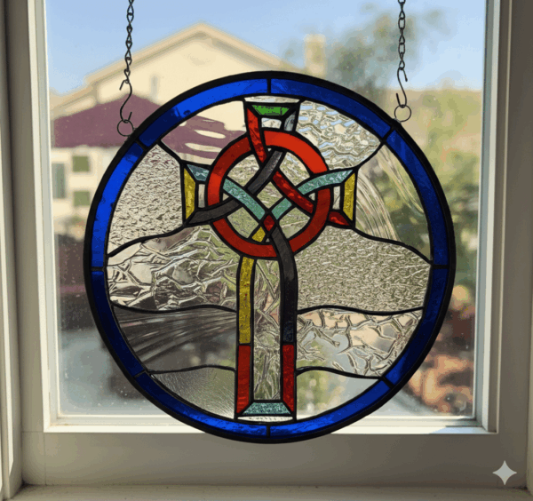 A round stained glass panel featuring a central Celtic knot cross in red, black, green, and yellow, surrounded by clear textured glass, with a deep blue glass border. It hangs in a sunlit window.