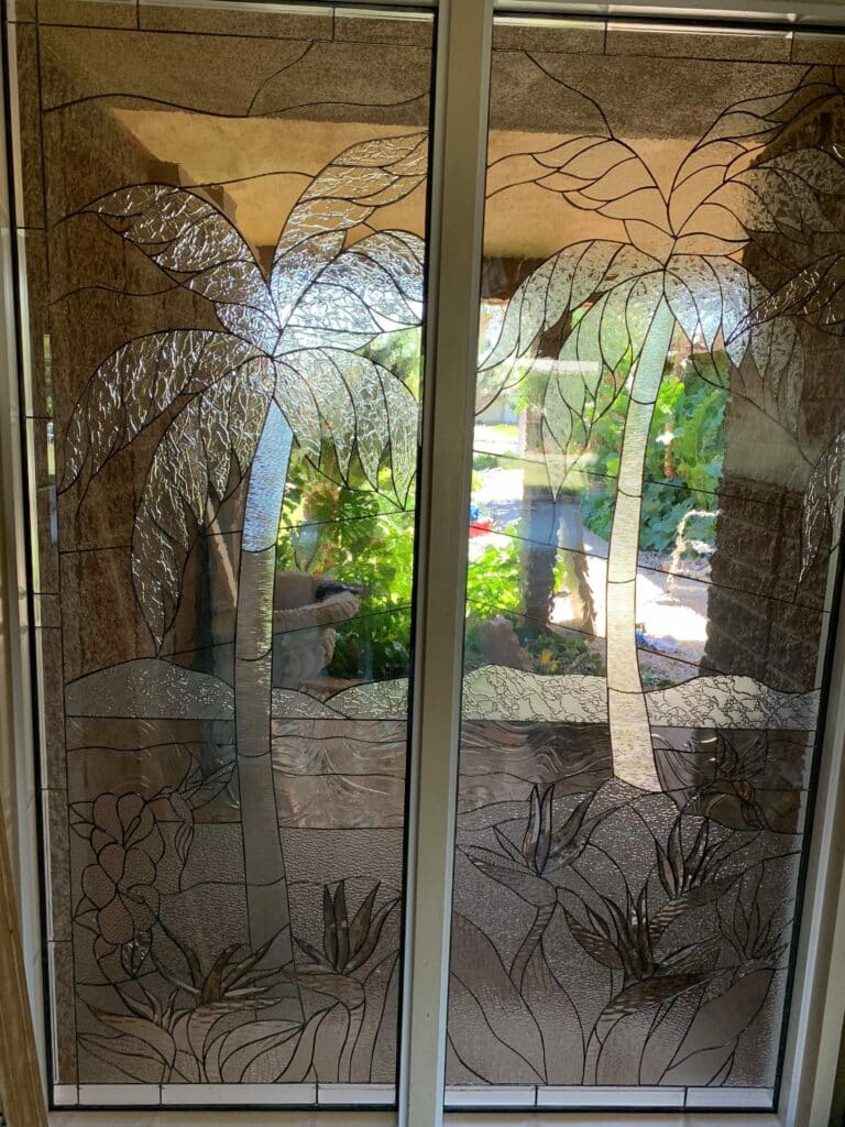 Two rectangular leaded stained glass panels forming a unified tropical scene. The design features two tall palm trees with textured fronds, and at the base, detailed Birds of Paradise flowers and foliage. The glass is predominantly clear with various textures including hammered, streaky, and reamy patterns, creating visual interest and privacy. Dark lead lines define all elements, set within light-colored window frames.