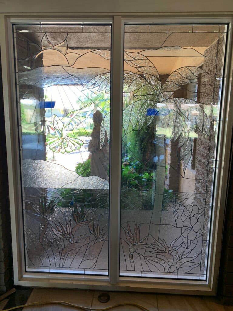 Two rectangular clear and textured stained glass panels in a white frame, depicting a tropical landscape with a soaring dove, sun rays, large leaves, and Bird of Paradise flowers.