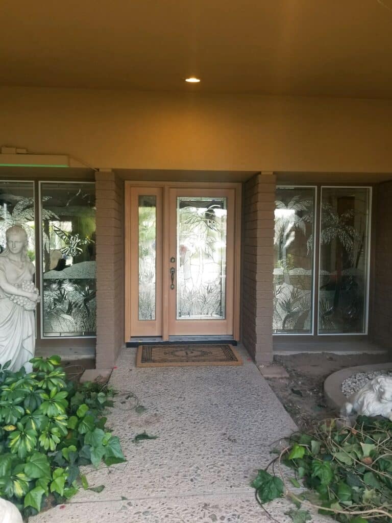 A front entryway with light wood double doors and two adjacent sidelights, all featuring clear textured glass panels with an intricate design of palm trees and tropical leaves.