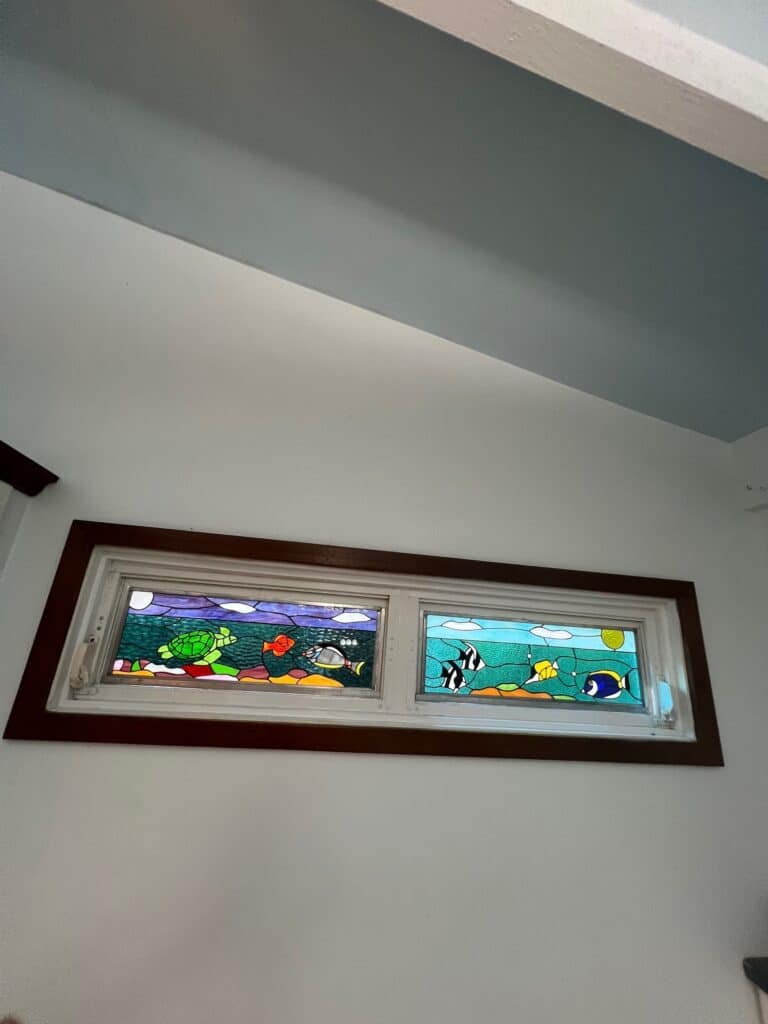 A rectangular, two-panel stained glass transom window depicting a colorful underwater scene. The left panel features a green sea turtle and various fish in purple and dark blue water. The right panel shows tropical fish, including striped ones, in light blue water.