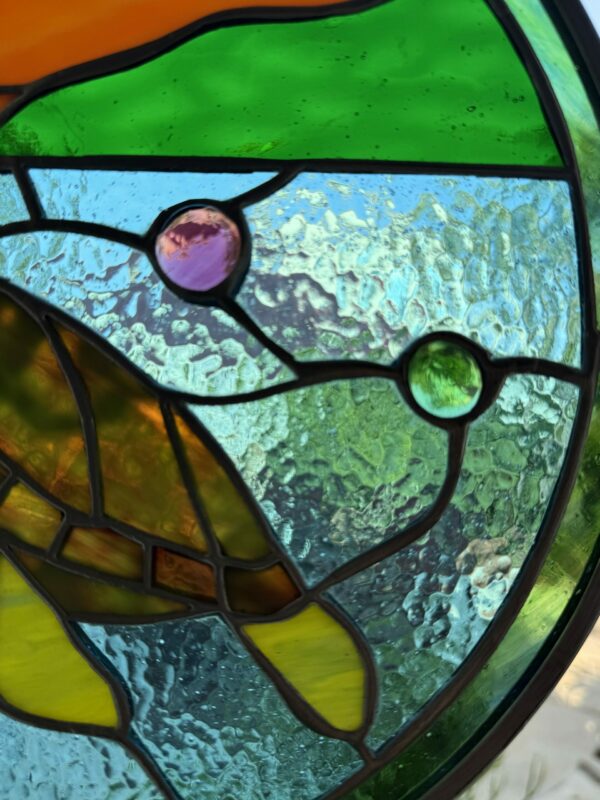 A close-up of an ornate stained glass panel featuring textured clear glass, green, blue, yellow, and orange segments, and two embedded round glass jewels in purple and green.