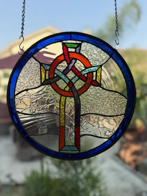 A round stained glass panel featuring a vibrant, interweaving Celtic knot cross in red, orange, green, and yellow, set against a background of clear textured glass and framed by a deep blue border. The panel hangs from a metal chain against an outdoor backdrop.