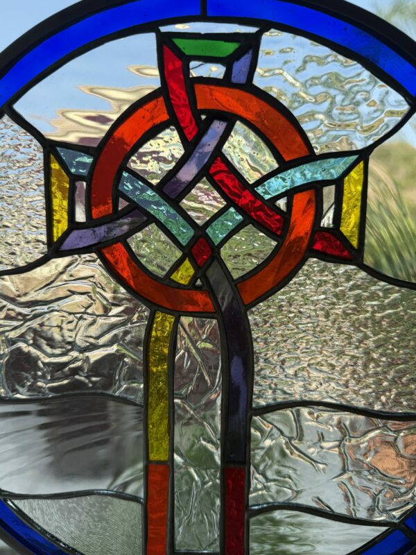 A circular stained glass panel featuring a multi-colored Celtic knot cross at its center. The knot is made of interwoven orange, red, blue, green, purple, and yellow glass, surrounded by various clear textured glass pieces, and framed by a top blue arc.