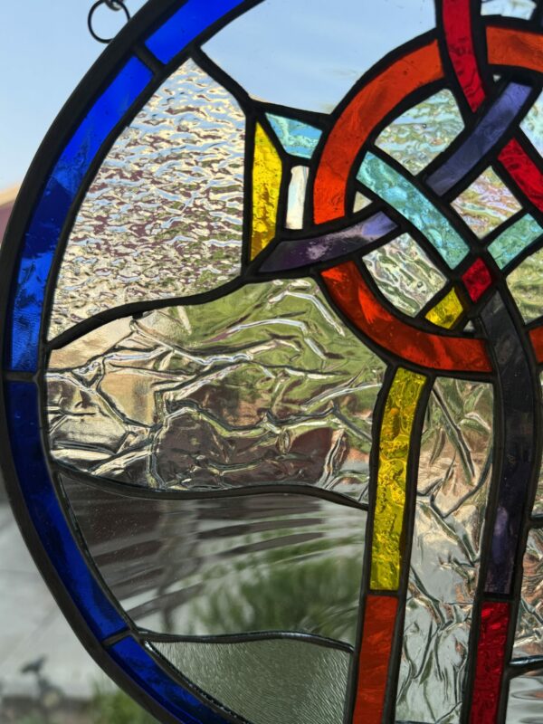 A close-up of a circular stained glass panel featuring a colorful, interwoven Celtic knot design with sections of blue, red, orange, yellow, purple, and various clear textured glass.
