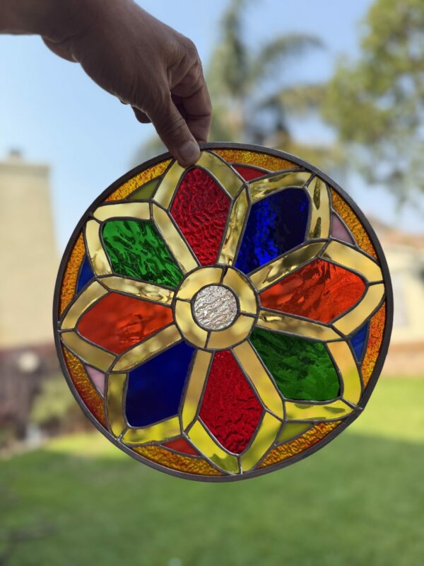 A hand holds a circular stained glass panel against an outdoor background. The panel features a vibrant, multi-colored geometric design resembling a flower or mandala, with textured clear, red, blue, green, and amber glass segments framed by lead lines.