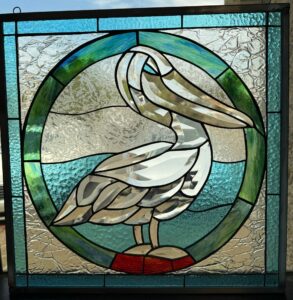 A rectangular stained glass panel with a pelican standing in profile, crafted from clear textured and beveled glass. The pelican is centered within a circular motif of blue and green glass representing water, set against clear patterned glass. The panel's corners are light blue textured glass, and it features a dark metal frame with a hanging hook.