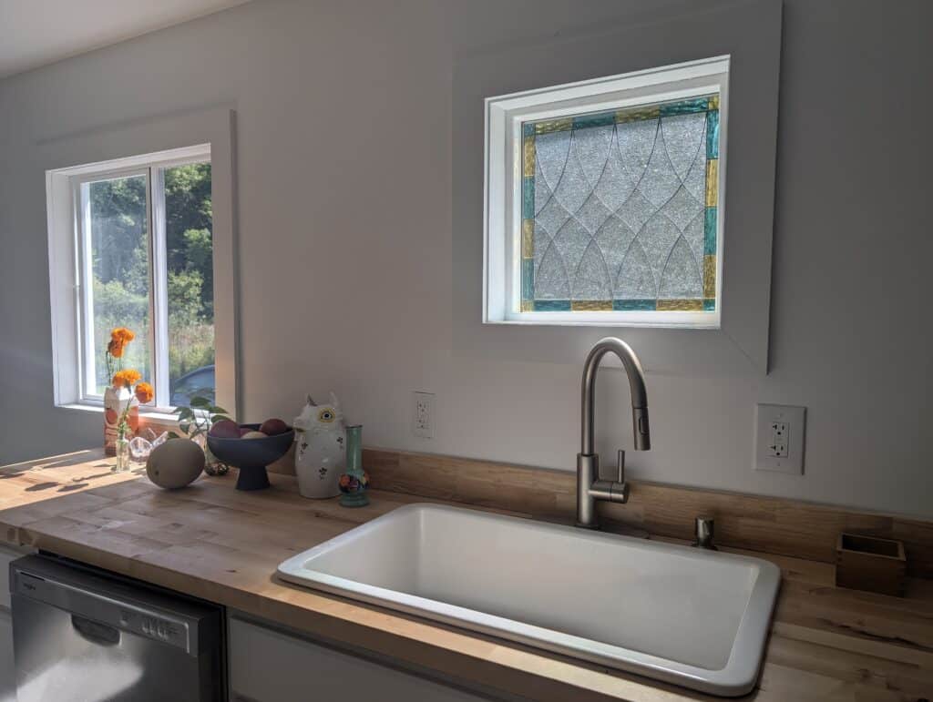 A rectangular stained glass window panel featuring clear textured glass in an arcing lattice pattern, accented with teal and amber border glass, installed in a bright kitchen above a sink.