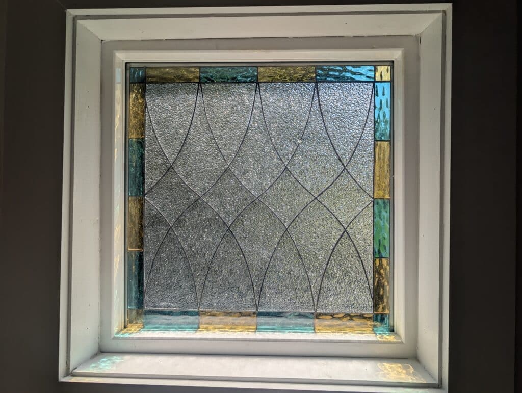 A square leaded stained glass panel with a clear, heavily textured center featuring an interlocking wave pattern, bordered by alternating rectangular pieces of golden yellow and blue-green glass. The panel is set in a white window frame.