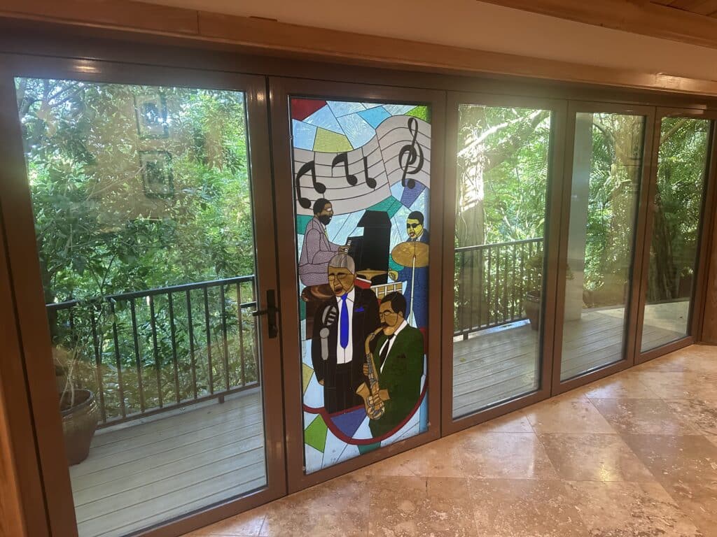 A rectangular stained glass panel, installed in a door, depicting a jazz band. Four musicians are shown: a pianist, a singer with a microphone, a drummer, and a saxophonist. Above them are musical notes on a staff. The background is a mosaic of abstract colored glass.