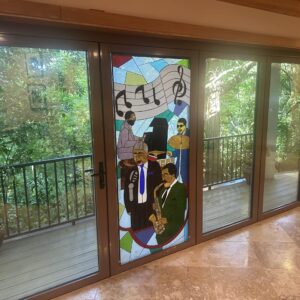 Rectangular Jazz Band Stained Glass Panel A rectangular stained glass panel, installed in a door, depicting a jazz band. Four musicians are shown: a pianist, a singer with a microphone, a drummer, and a saxophonist. Above them are musical notes on a staff. The background is a mosaic of abstract colored glass.