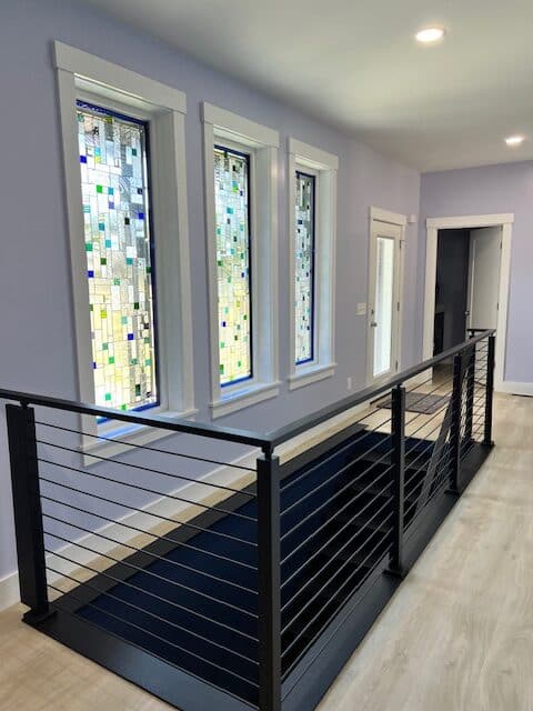 Three tall, rectangular stained glass windows featuring an abstract mosaic design with clear textured glass and scattered blue, green, and yellow square accents, installed in a modern hallway.