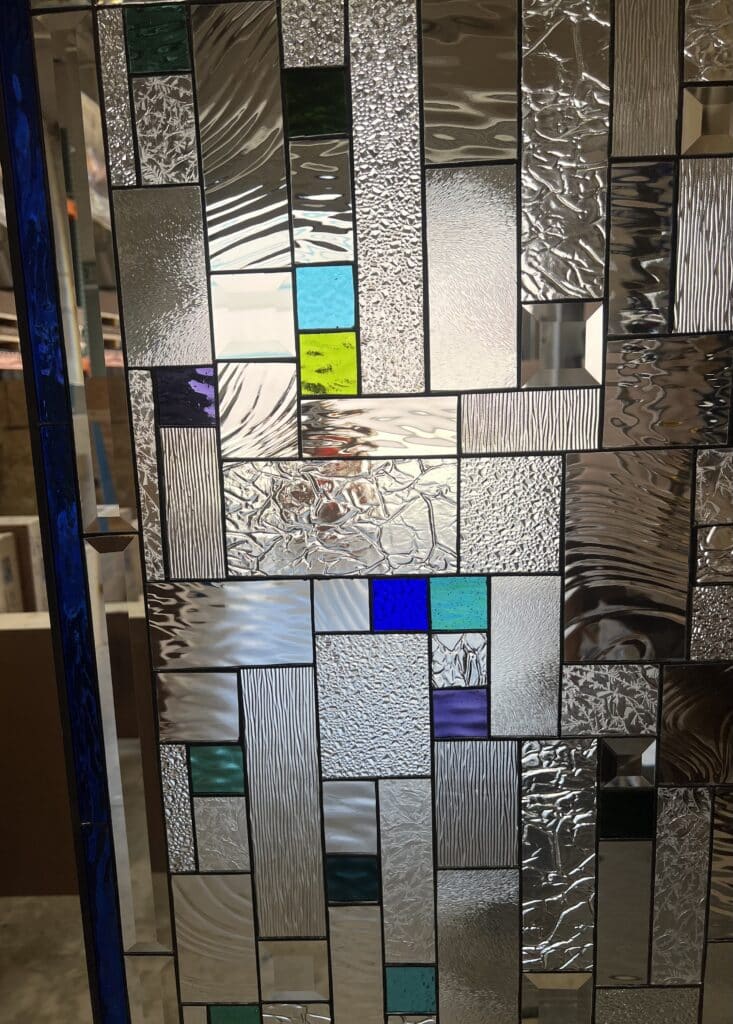 A rectangular, leaded stained glass panel with an abstract geometric design, featuring various clear textured glass patterns and small colored squares in blue, green, and purple.