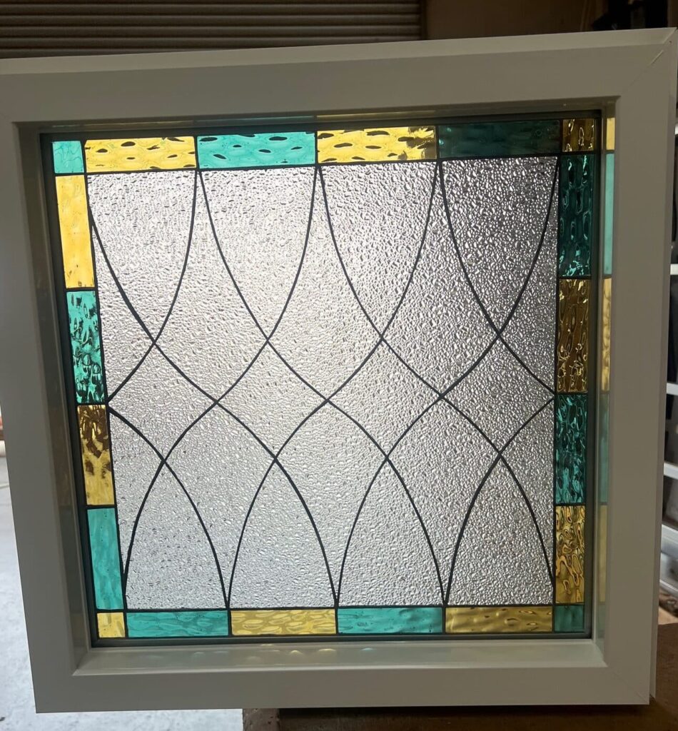 A rectangular white-framed insulated window featuring a stained glass panel. The panel's center is clear textured glass with a flowing geometric pattern created by black lead lines forming intersecting arches. A border of alternating rectangular pieces of bright yellow/amber and teal stained glass surrounds the clear center.
