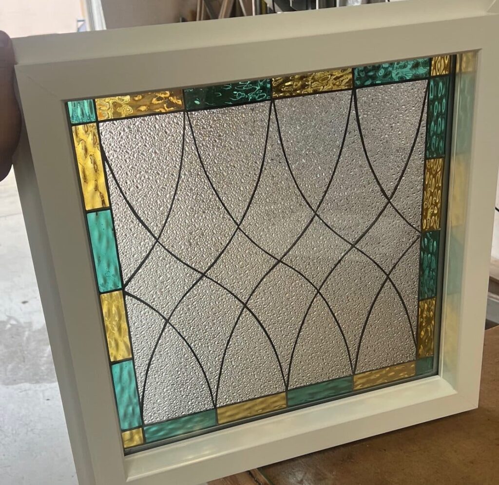 A square stained glass insulated window in a white vinyl frame, featuring a clear textured glass center with an intersecting geometric leaded pattern, bordered by alternating gold and teal rectangular glass pieces. A person's hand holds the window in a workshop.