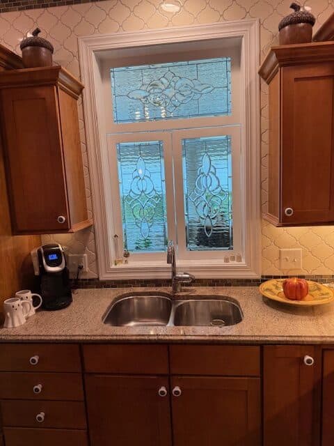 A multi-panel rectangular, leaded clear beveled stained glass window with ornate scrollwork designs, installed above a kitchen sink.