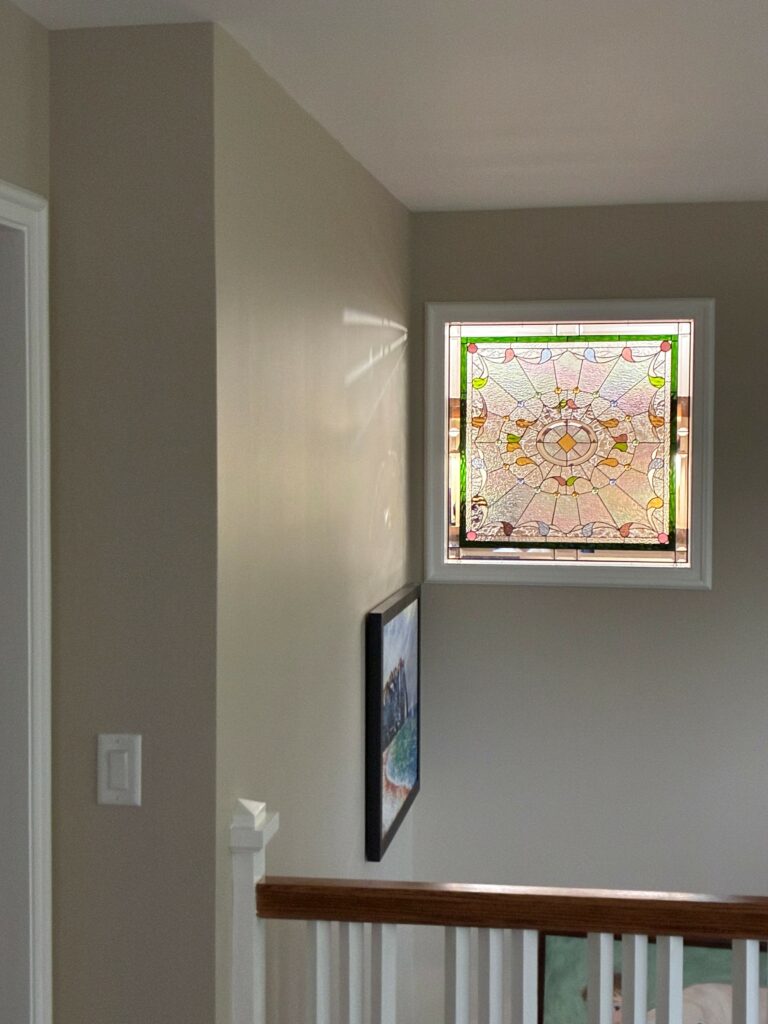 A square, white-framed, leaded stained glass window with a circular medallion at its center, surrounded by radiating clear textured glass and colorful petal-like details in a neutral-toned hallway.