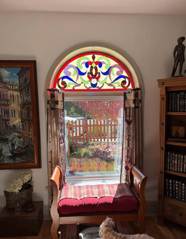 An arched, leaded stained glass panel featuring a symmetrical ornate scroll design in red, blue, green, and purple, installed above a rectangular window.