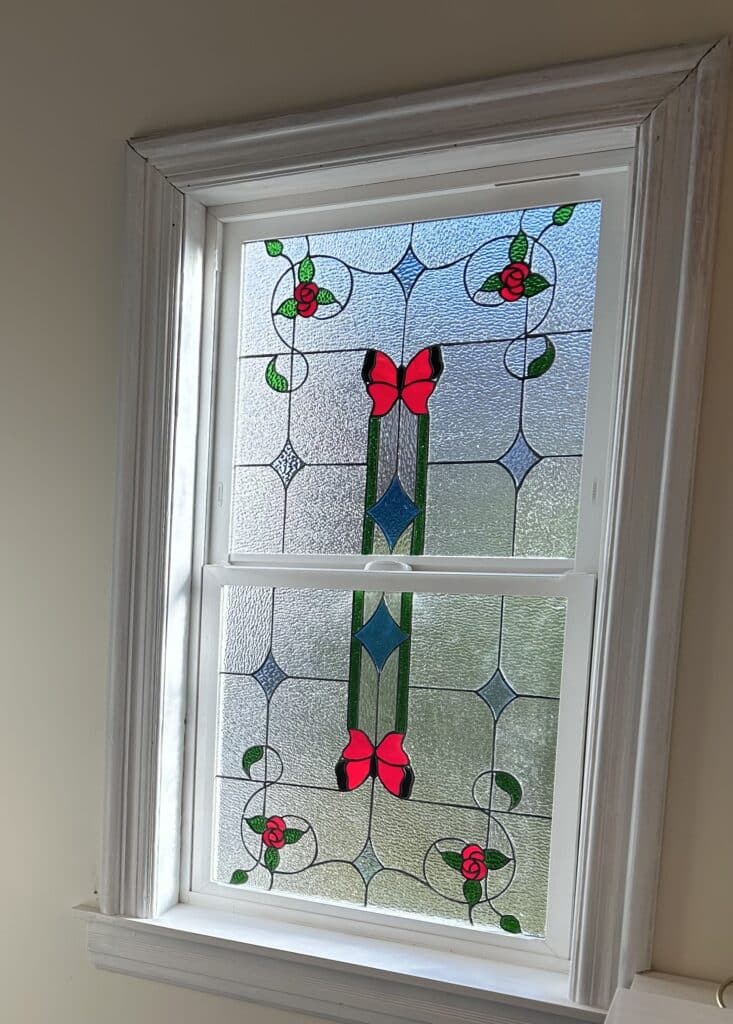 A rectangular double-hung window featuring an integrated stained glass panel with symmetrical red butterflies, red roses, green leaves, and clear textured glass.