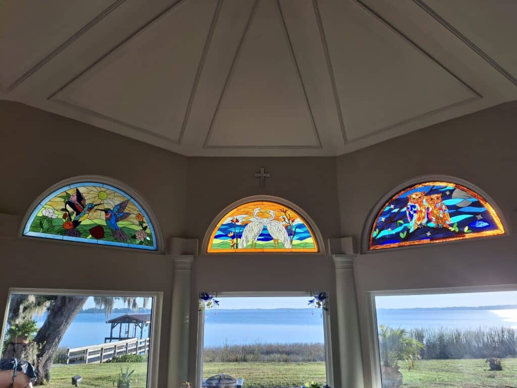 Three arched stained glass windows depicting nature scenes, installed above clear windows with a lake view. The left panel shows two hummingbirds among flowers; the middle panel features two egrets in reeds; and the right panel illustrates two owls on a branch under a starry night sky.