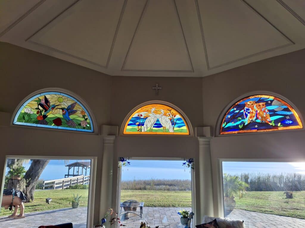 A set of three arched stained glass transom windows. From left to right, the panels depict: two hummingbirds with flowers, two white cranes, and two owls on a branch at night.